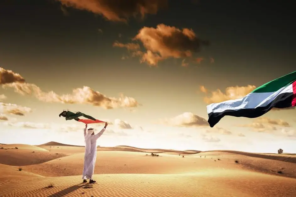 A man is standing in the desert holding a uae flag.
